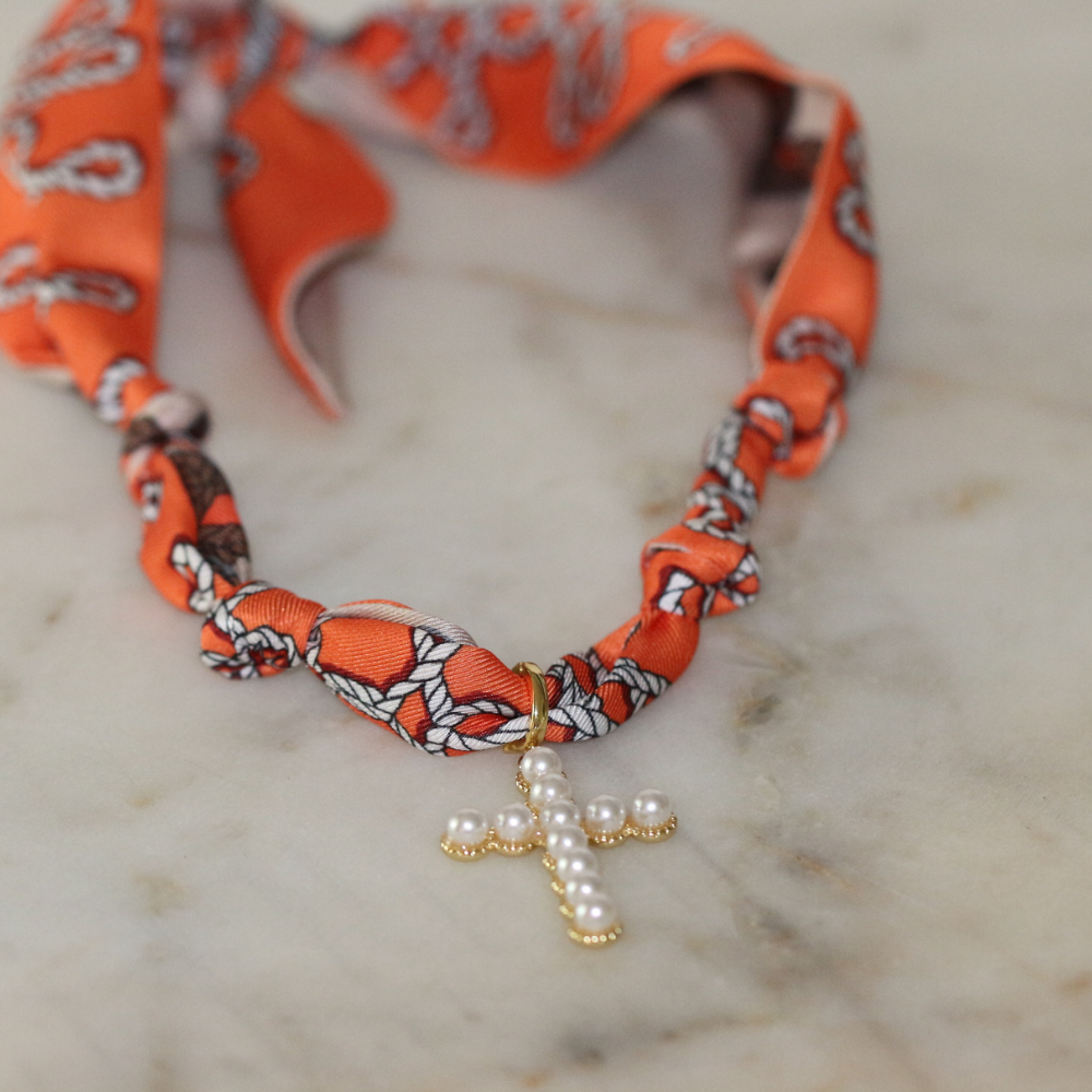 Necklace with orange fabric strap and pearl cross pendant on a marble surface